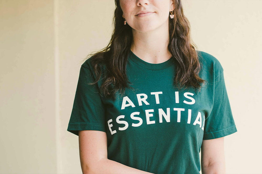 Dark green t-shirt with bold white text 'ART IS ESSENTIAL' printed, worn by a person against a plain background.