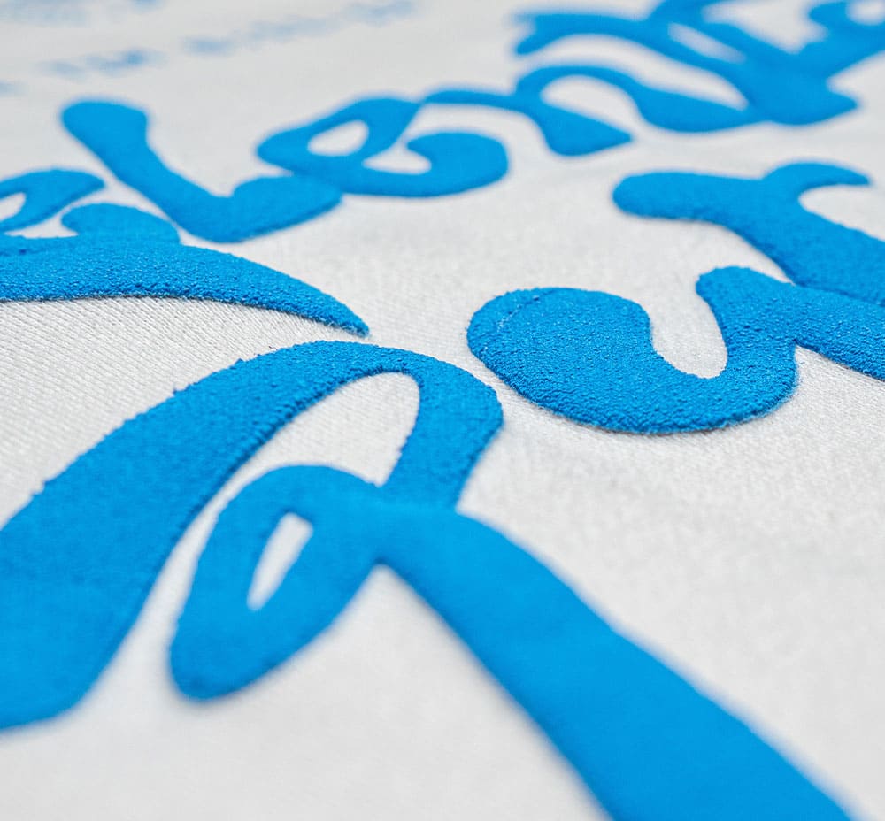 Screen Printing - Puff Printing Close-up of textured blue puff print text on a white background, showcasing a three-dimensional effect.