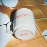An off-white ceramic stacking diner mug featuring a custom red logo for a surf shop on the side and a matching custom printed logo on the bottom base, resting on a tiled surface.